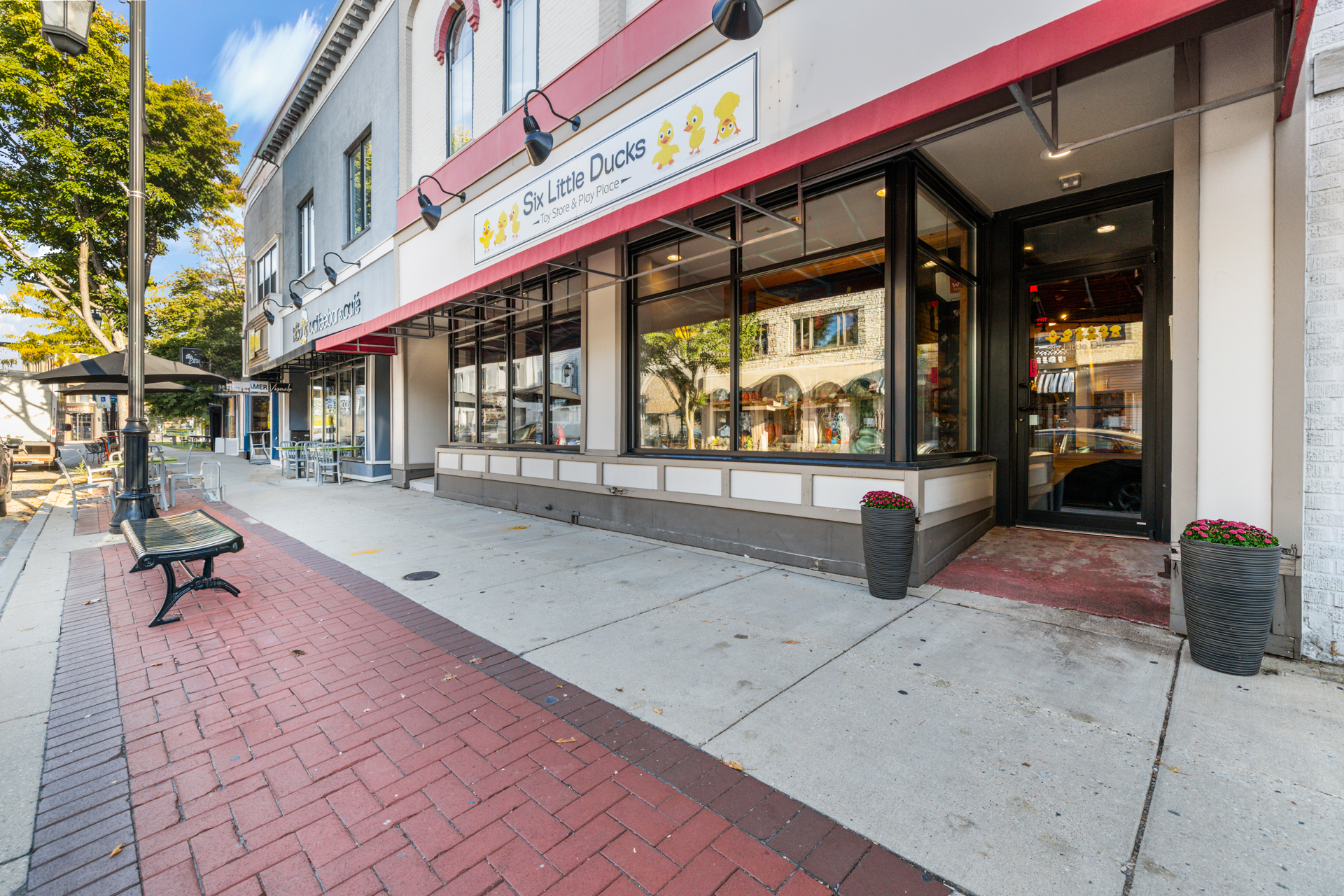 The Six Little Ducks Toy Store & Play Place storefront on E Wisconsin Ave in downtown Oconomowoc, with large glass windows, a white and red sign with six yellow ducks, and a black bench on the red brick sidewalk