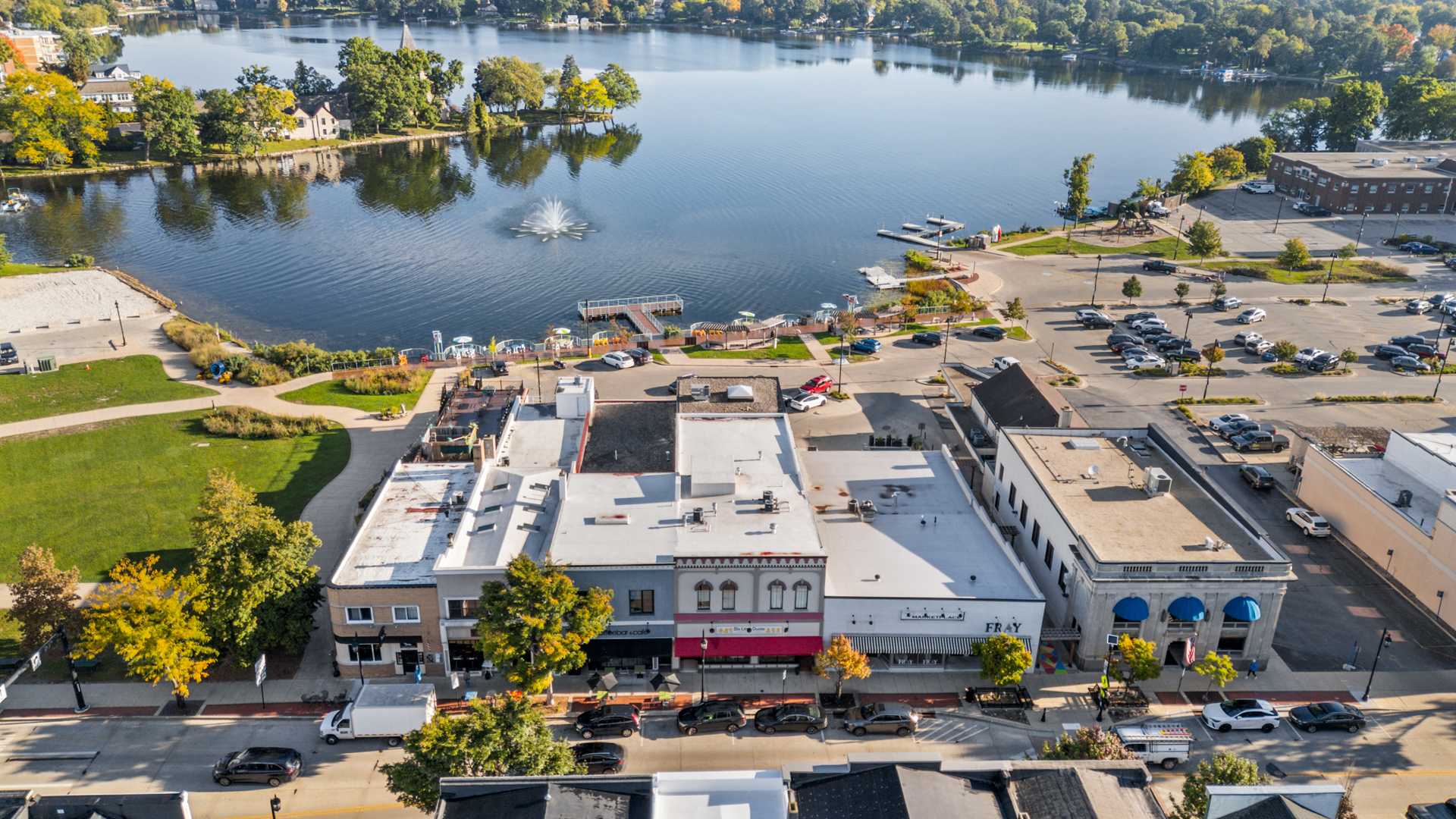 A wide aerial view of downtown Oconomowoc, with a row of storefronts including Six Little Ducks below a green park and a large blue lake with a fountain in the middle
