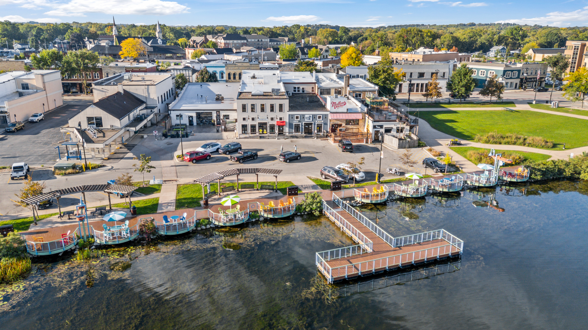 An aerial view of downtown Oconomowoc showing the Six Little Ducks building behind a waterfront park, with a T-shaped dock and circular seating pods holding colorful chairs out on the blue lake