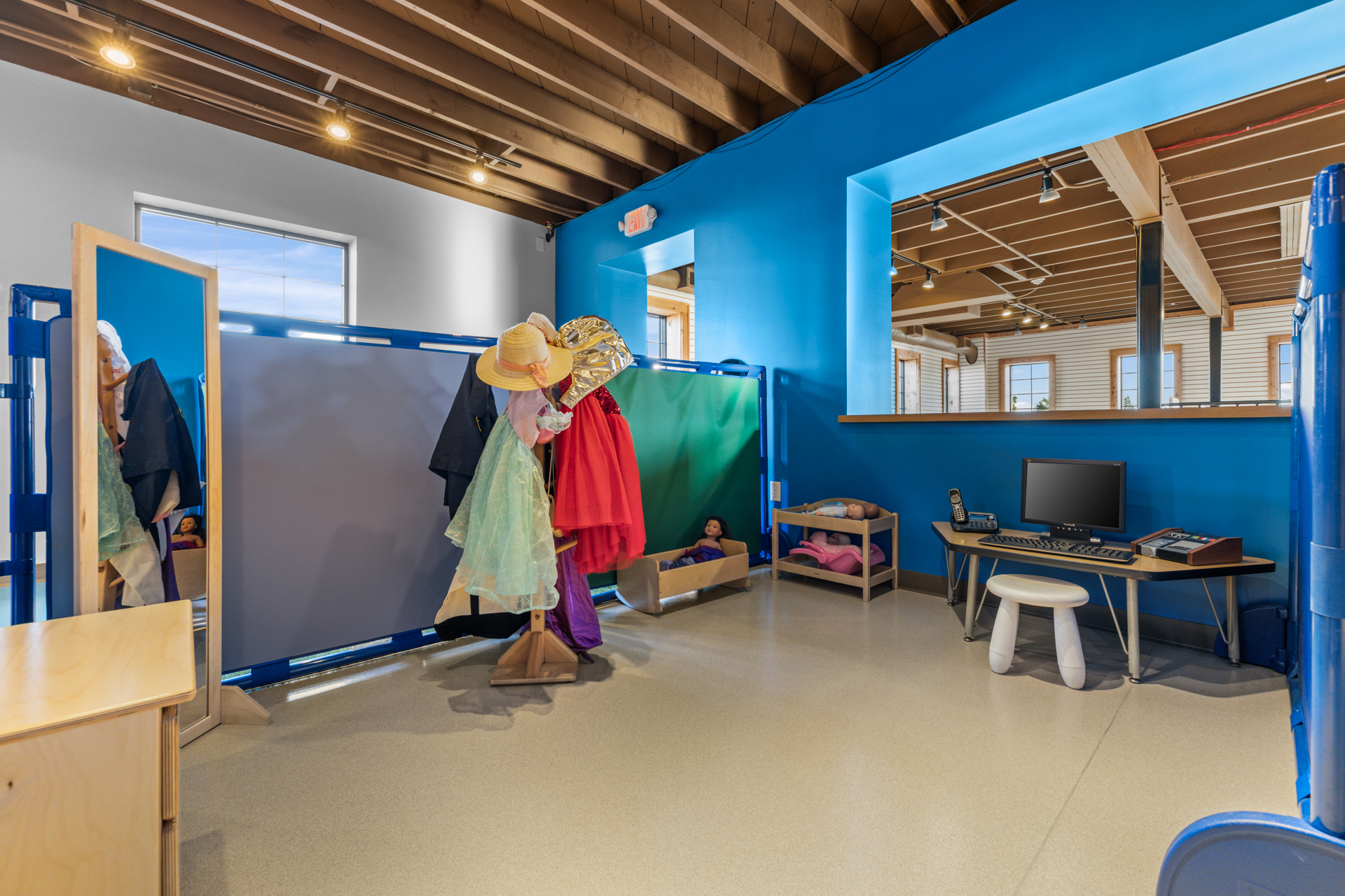 Dress-up area with a costume rack, tall mirror, doll bed and pretend office desk on a bright blue wall at Six Little Ducks